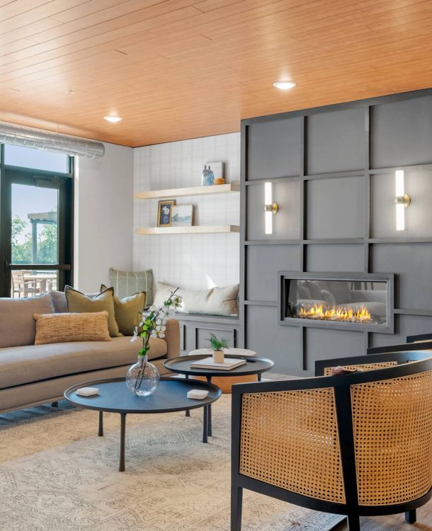 Lexi Apartments Modern living room with a gray fireplace, beige sofa, two woven chairs, and large windows letting in light.