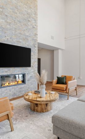 Lexi Apartments Modern living room with a stone fireplace, TV, tan chairs, gray sofa, and large windows with outdoor view.