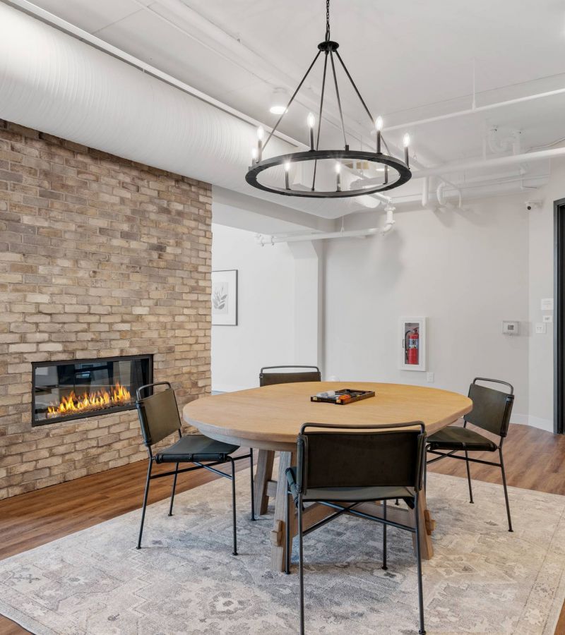Lexi Apartments Modern meeting room with round table, four chairs, brick fireplace, and large windows letting in natural light.